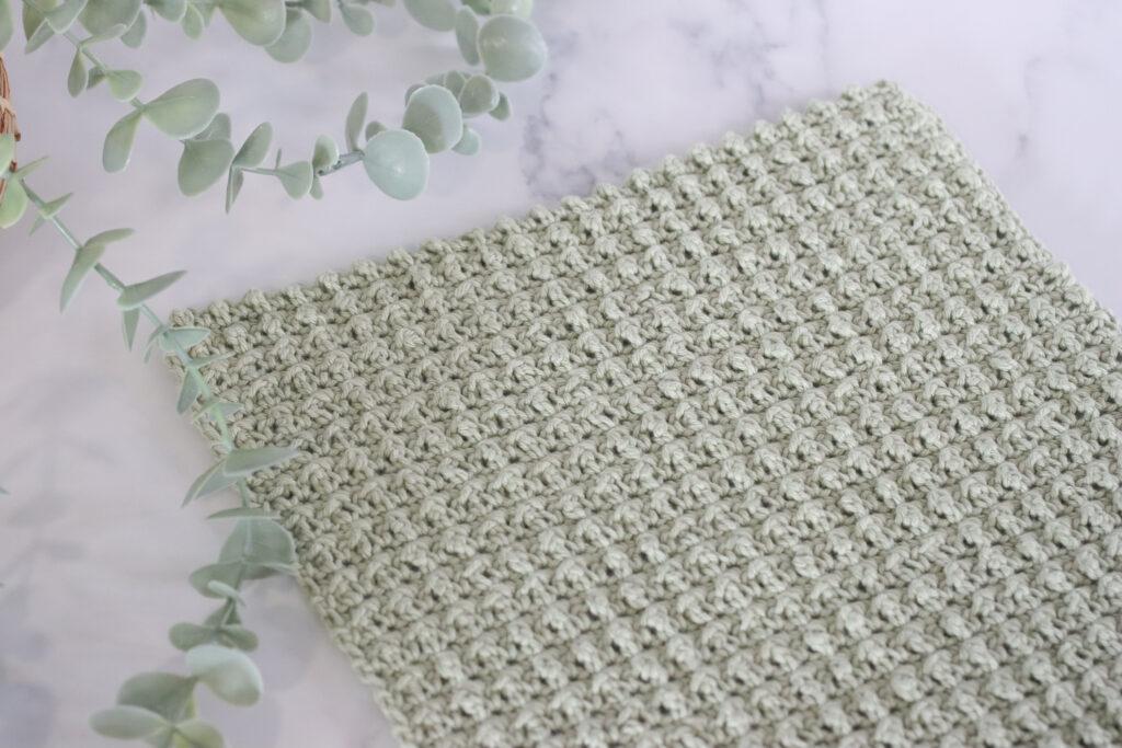 Close-up of a crocheted swatch using the cobble stitch in light sage green yarn, displayed on a marble surface with eucalyptus leaves in the background.