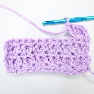 An in-progress swatch of popcorn stitch sits on a white background. The swatch is made from lilac yarn and uses a metallic blue crochet hook.