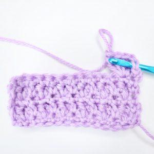 An in-progress swatch of popcorn stitch sits on a white background. The swatch is made from lilac yarn and uses a metallic blue crochet hook.