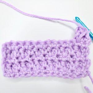 An in-progress swatch of popcorn stitch sits on a white background. The swatch is made from lilac yarn and uses a metallic blue crochet hook.