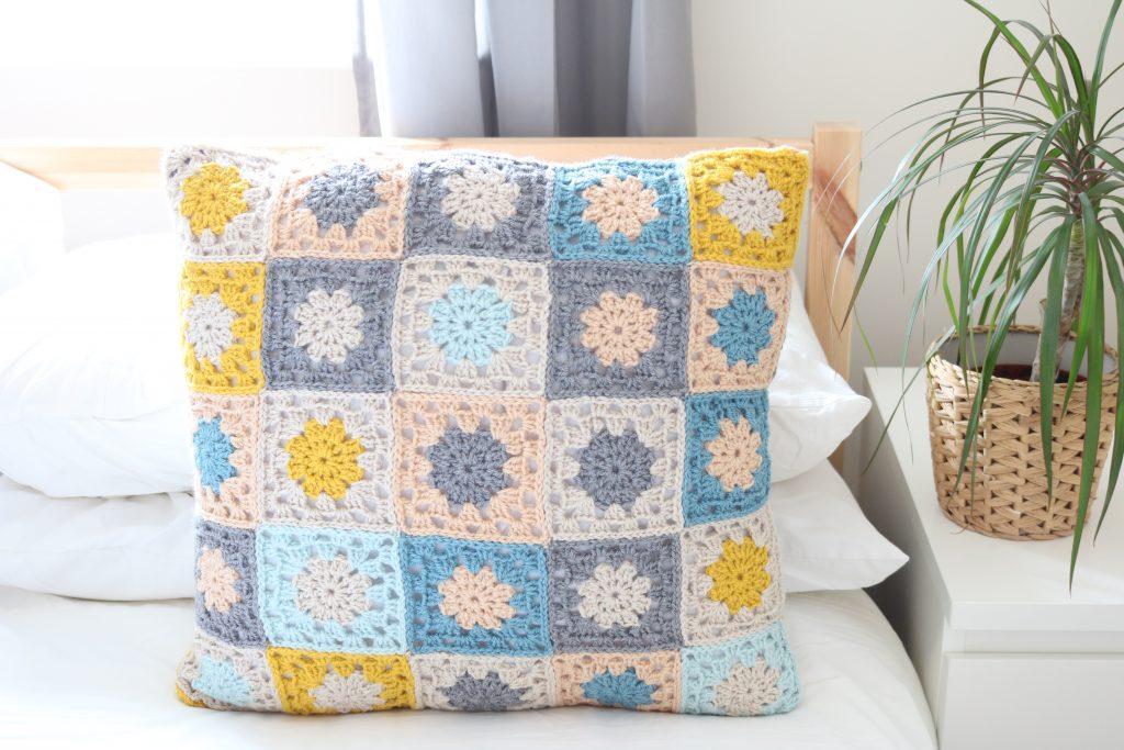 a granny square cushion made of mustard yellow, grey, pale blue and peach yarn sat on a white bed with a wooden frame. Next to the bed is a white bedside table with a plant sat in a wicker pot.