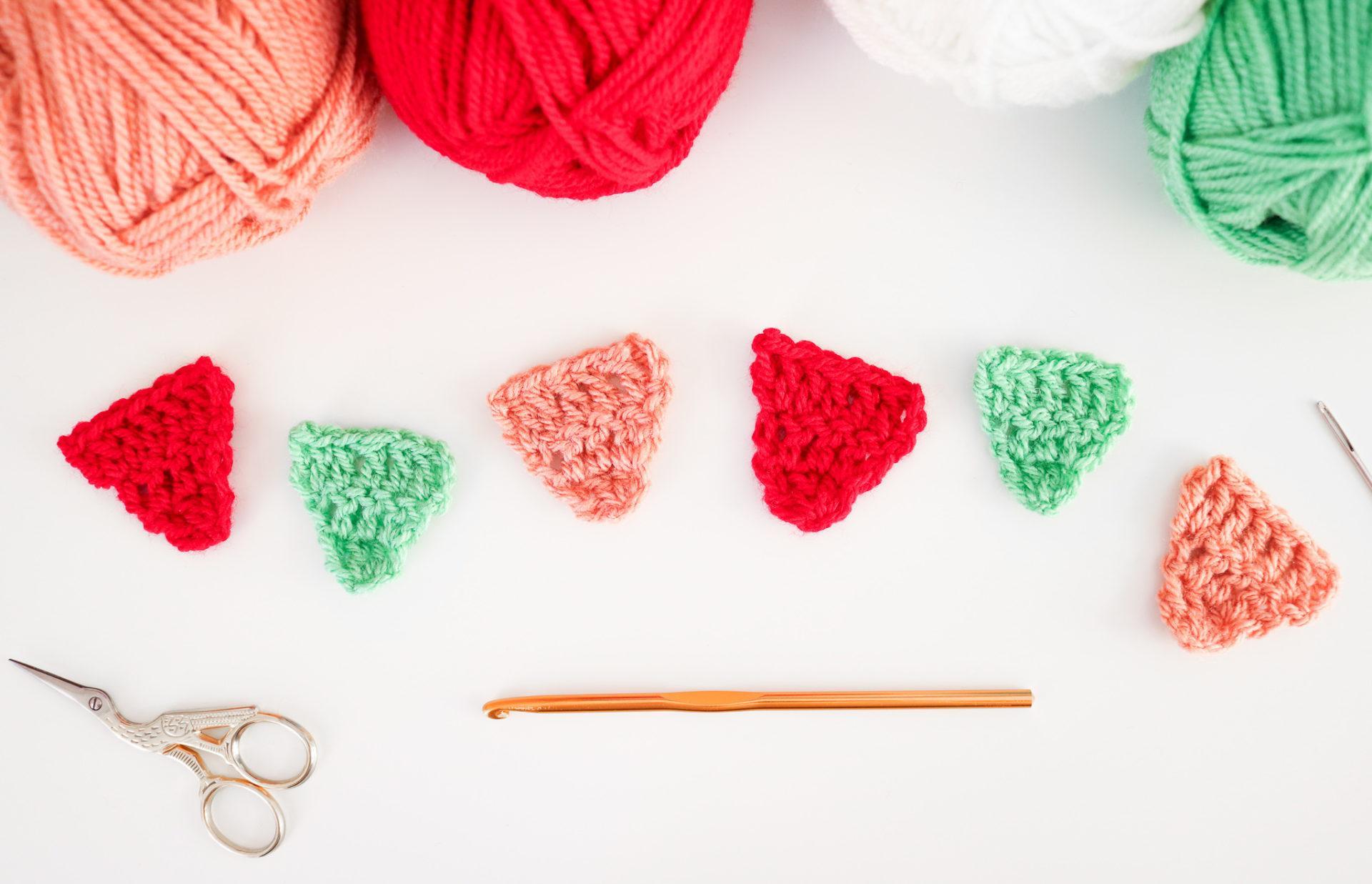 a flat lay image of orange, red, white and green yarn with mini bunting triangles lay under the yarn with a crochet hook and scissors on a white background.
