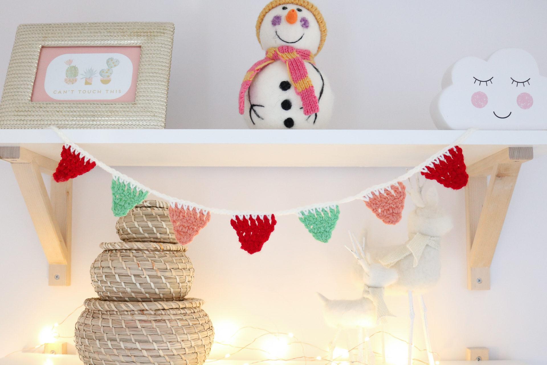 mini crochet bunting hung from a shelf in a bedroom, the shelves have some baskets, a ceramic cloud, a photo frame and a felt snowman on them.