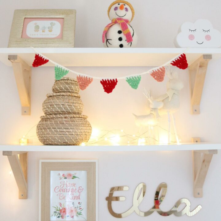 mini crochet bunting hanging from a white shefl in a little girls bedroom, the helfs are decorated with christmas ornaments and photo frames. The bunting is made in red, green and peach yarn attached together with cream yarn.