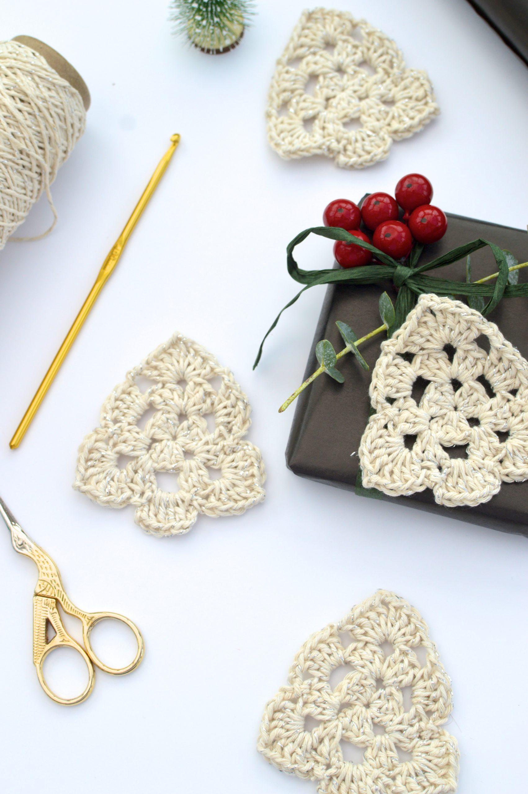 flay lay image of four crochet Christmas trees made in cream yarn lay on a white backdrop with gold scissors, gold crochet hook, yarn and a wrapped gift showing the crochet Christmas tree as a present topper along with foliage and berries. 