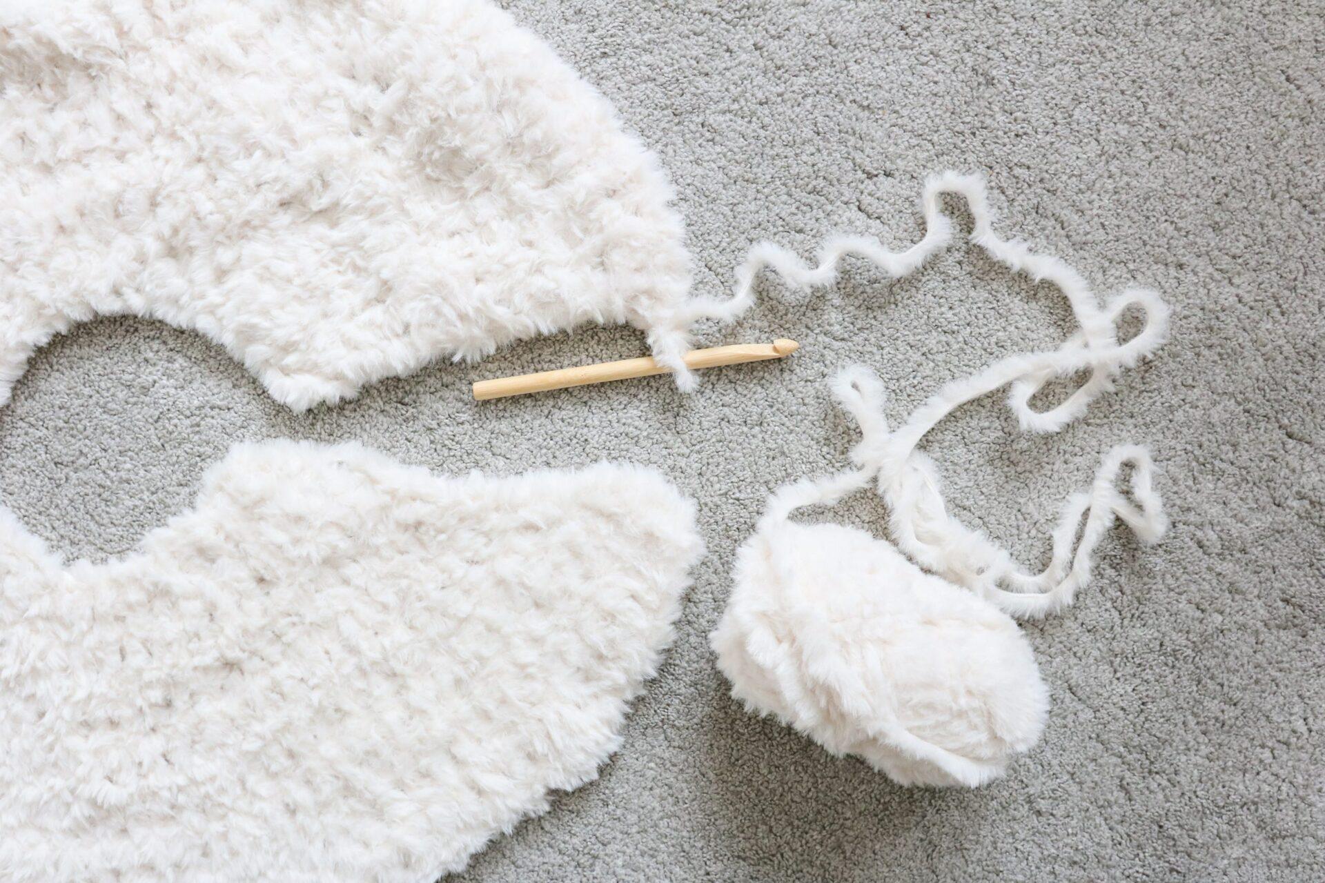a flat lay image of a crochet tree skirt made from cream fluffy yarn lay on a beige carpet with the ball of yarn still attached and a wooden crochet hook.