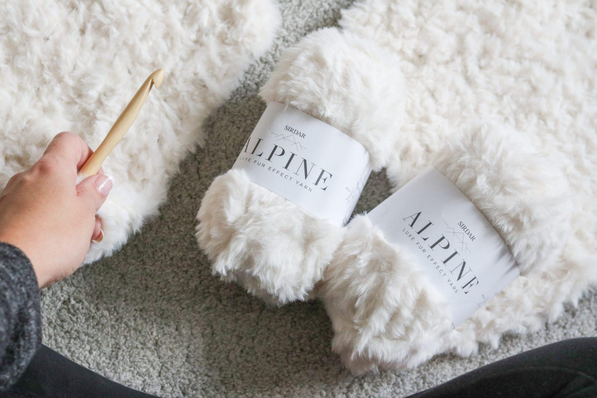 two balls of sirdar alpine yarn in cream lay on beige carpet with a hand holding a wooden crochet hook to the left of the image.