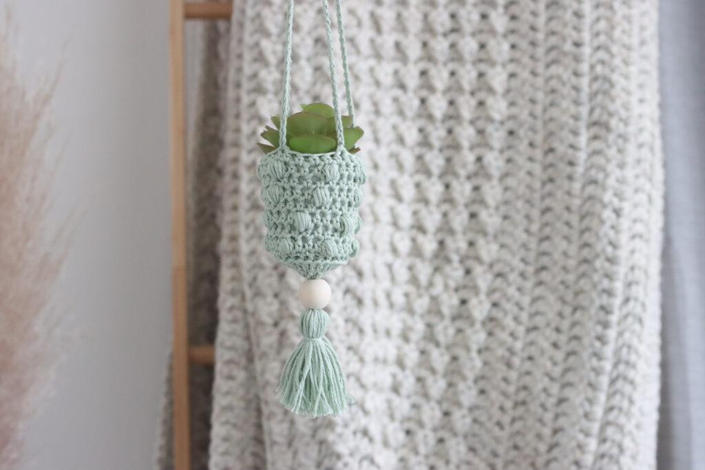 a mini crochet hanging plant holder made in pale green yarn. Hanging in front of a cream crochet blanket.