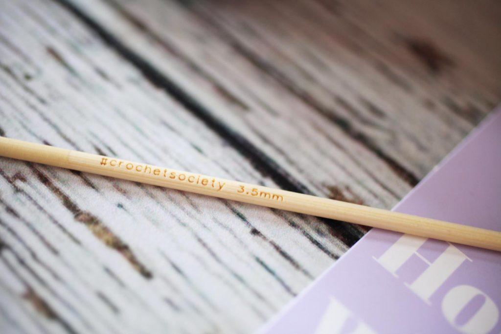 A bamboo crochet hook with #crochetsociety sits on a pale wooden background.