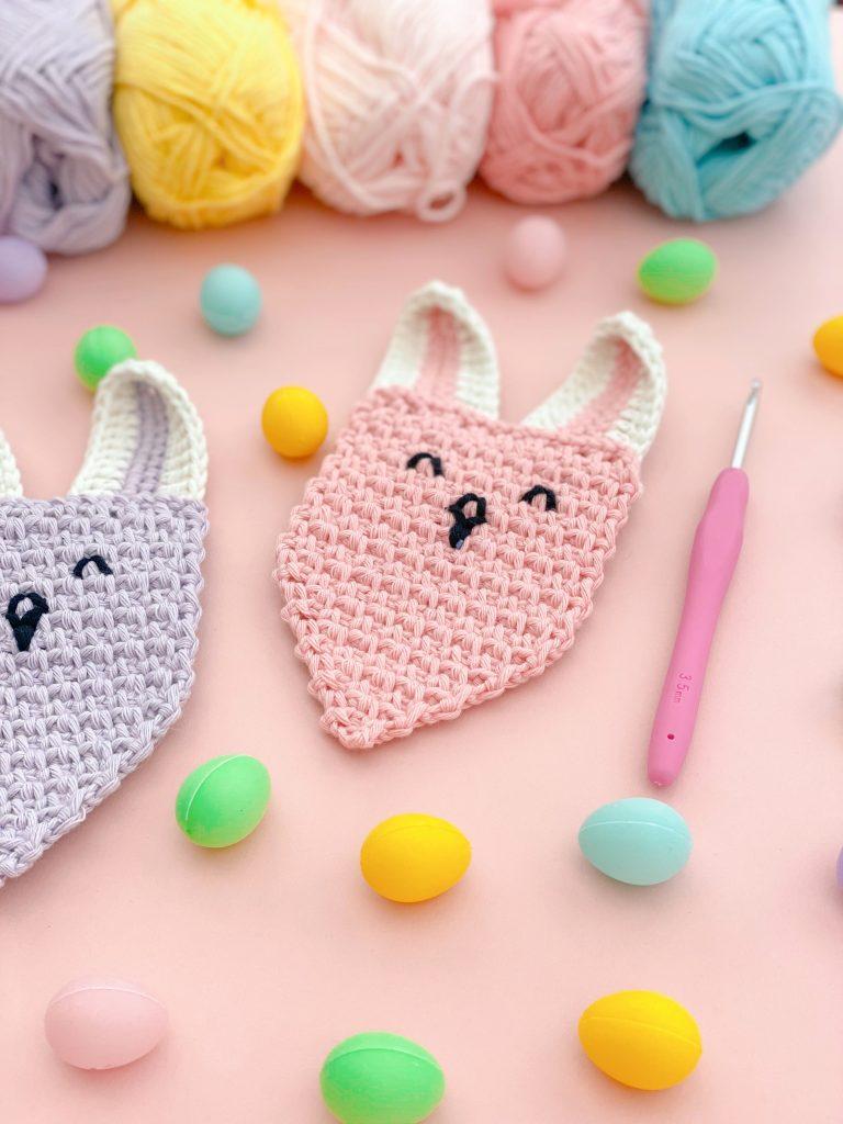 A pair of flags from crocheted bunny bunting sit beside a pink crochet hook and pastel coloured easter eggs.