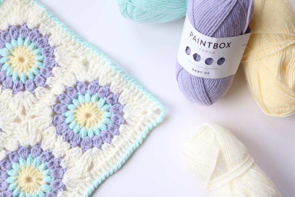 Sunburst Granny Square on a table with some purple Paintbox yarn and some other yellow, aqua and ivory yarn.