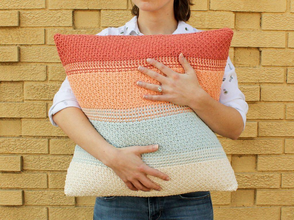 A woman holds a crocheted pillow in warm Summer colours. She stands against a yellow brick wall.