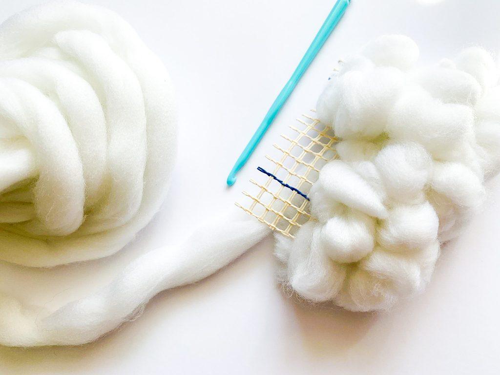 White wool intertwined into a large mesh with a blue plastic crochet hook to make a 3D cloud.