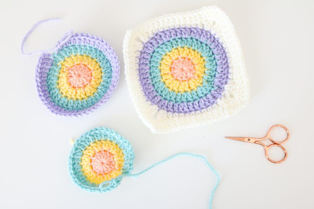 3 Circles of Hope granny squares in progress, one with 3 rounds, one with 4 rounds and one completed with the 5th round making it a square. A pair of rose gold scissors lays beside them.