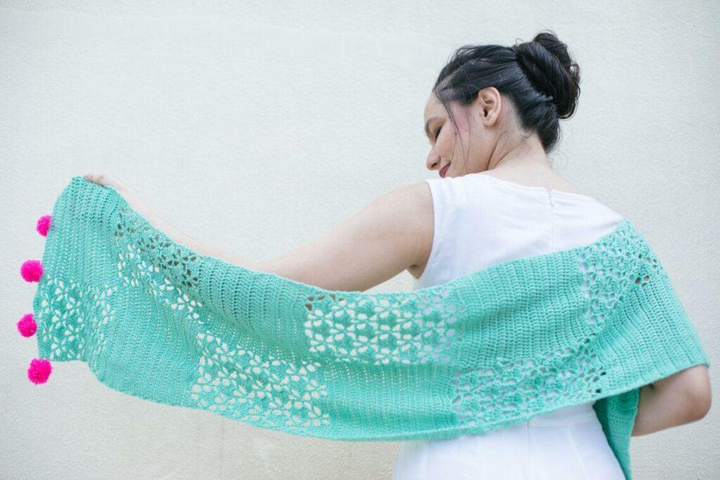 A lady holding a shawl that is sear foam in colour with pink pom pom ends.