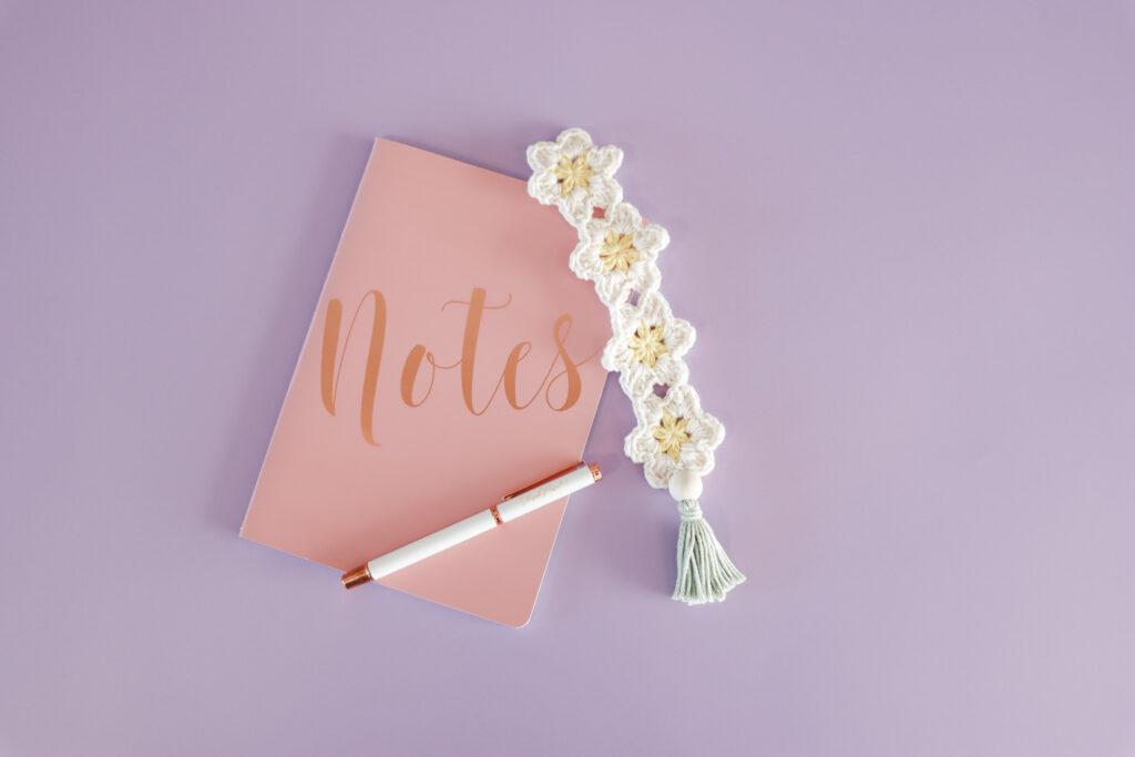 crochet daisy bookmark lay on a purple backdrop with a pink notebook that says notes across the cover in rose gold writing and a white and rose gold pen is lay across the note book.