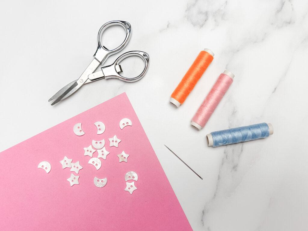 flay lay of three embroidery yarn spools in three colours; orange, pink and blue, sewing needle, scissors and white moon and star shaped beads on marble backdrop, the beads are lay on a pink piece of paper.