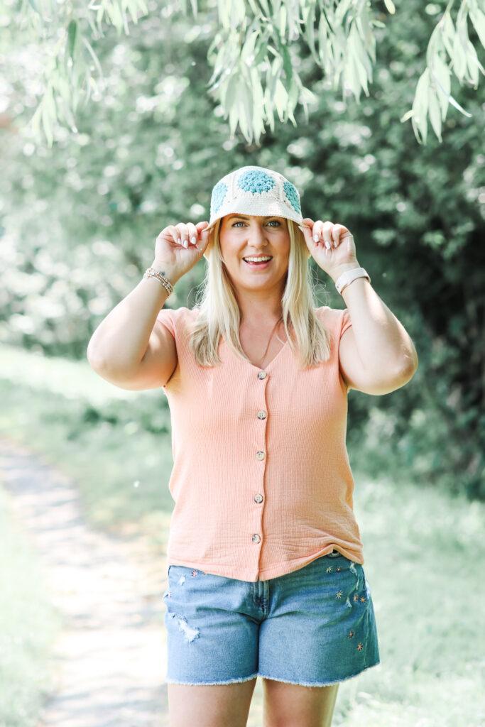 female stood outside under a tree wearing a peach buttoned t-shirt and denim shorts wearing a crochet granny square bucket hat. female is holding either side of the bucket hat smiling at the camera.