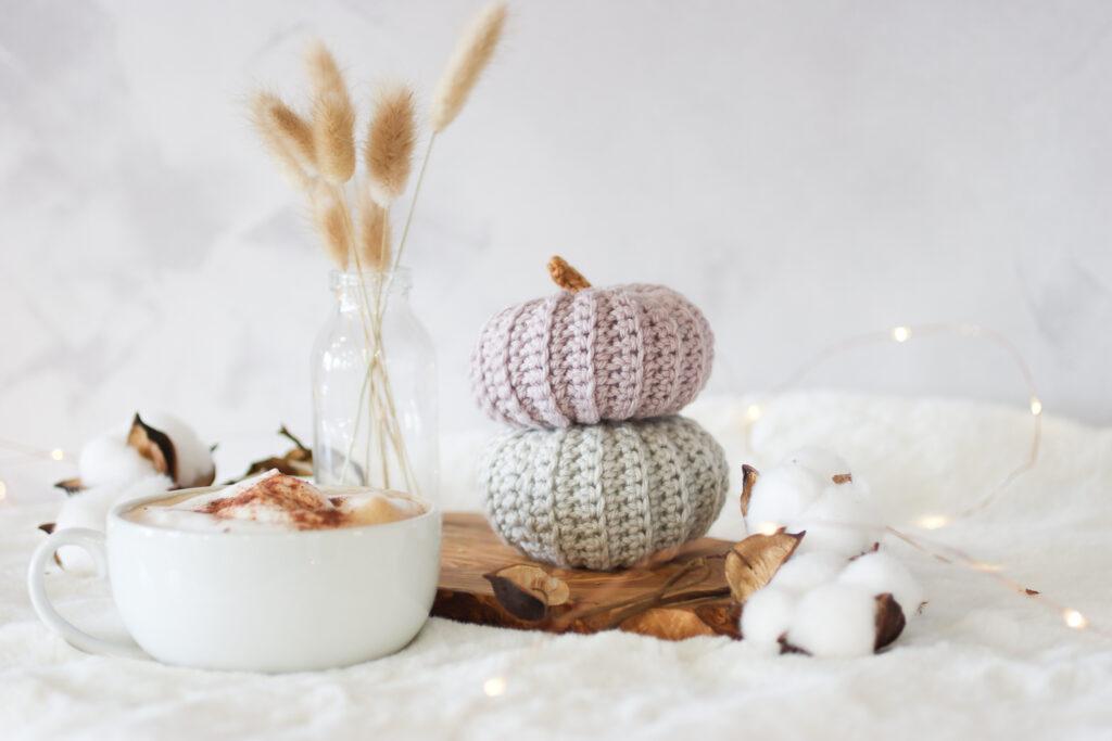 two crochet pumpkins stacked one grey and the other in mushroom. The crochet pumpkins are surrounded by small fairy lights with a cup of coffee and bunny tail dried flowers in a small glass vase. the background is white and the set up is placed on a white faux fur rug