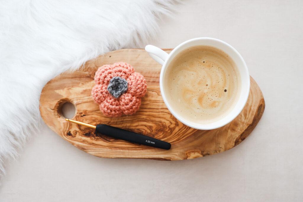 a flat lay image of a crochet pumpkin made in burnt orange yarn lay next to a cup of coffee on a wooden slice with a black and gold crochet hook
