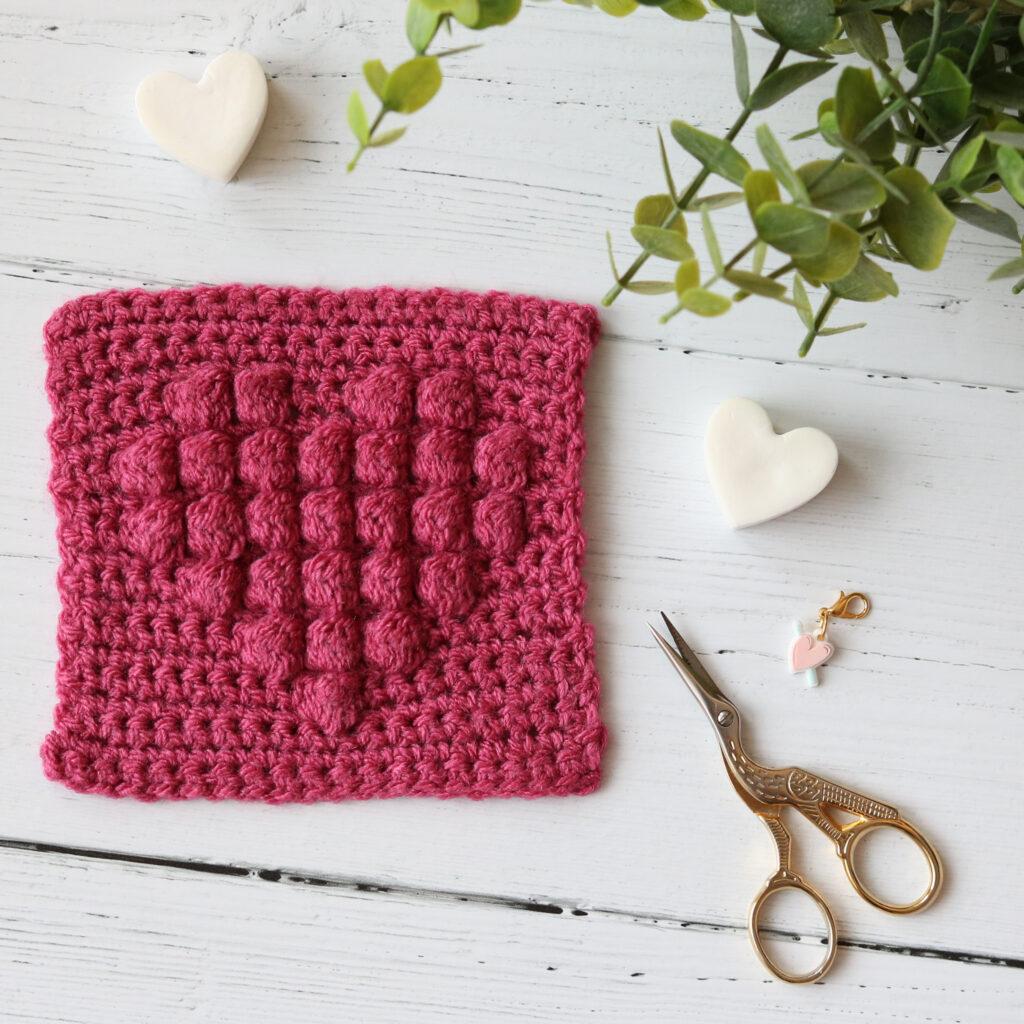 A swatch of crochet in reddish-pink with a bobble heart motif sits alongside scissors and a heart-shaped stitch marker.