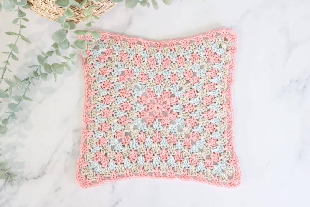 a flay lay of a spiked granny square made in pastel pink, blue and cream yarn lay on a marbled backdrop with foliage to the left of the swatch.