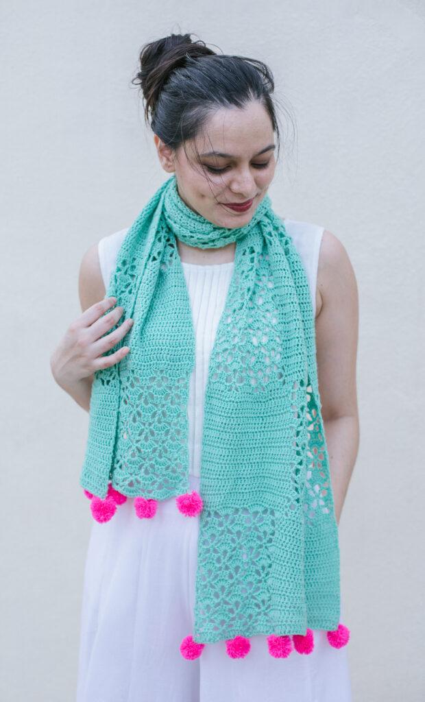 A woman is wearing a white dress and a seafoam-coloured crochet shawl with panels of lace and hot pink pompoms on the end. She is smiling.
