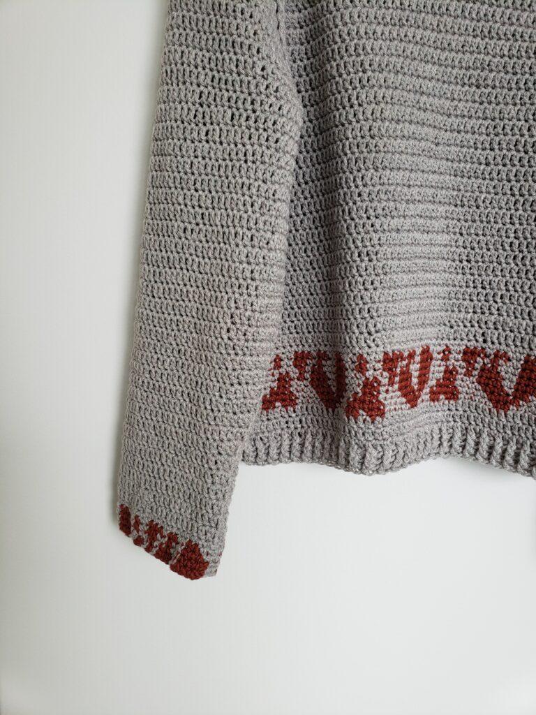 A taupe crochet sweater with a deep red animal trim motif on the bottom hangs up against a white wall.