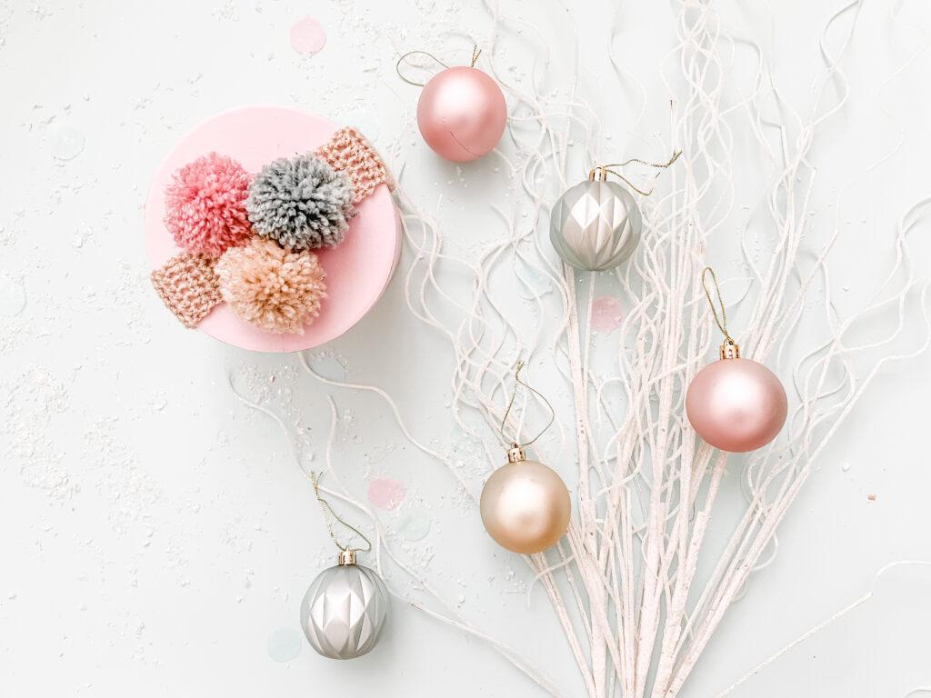 a flay lay image of a pink box with crochet ribbon wrapped around it decorated with three pom poms made in gold, silver and pink yarn. lay alongside the gift is silver sparkly branches and pastel coloured baubles.