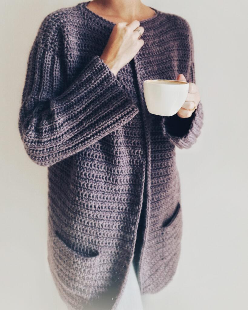 A woman wears a deep purple crochet cardigan and grey jeans. She is standing against a white background.