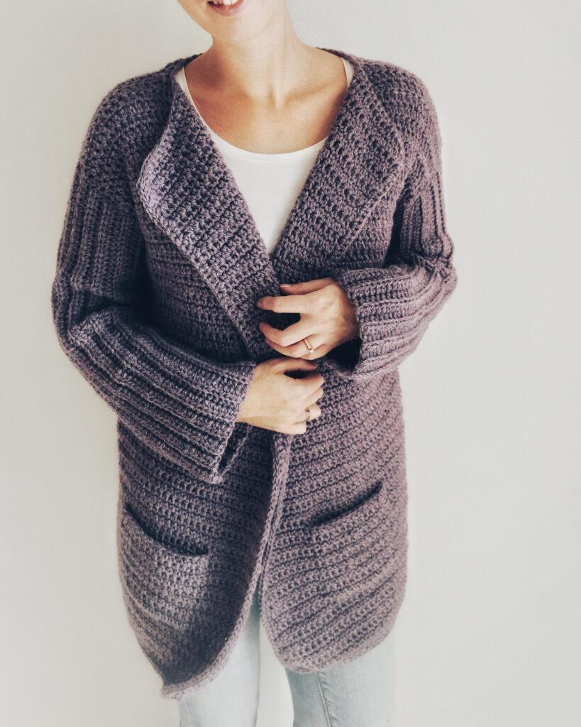 A woman wears a deep purple crochet cardigan and grey jeans. She is standing against a white background.