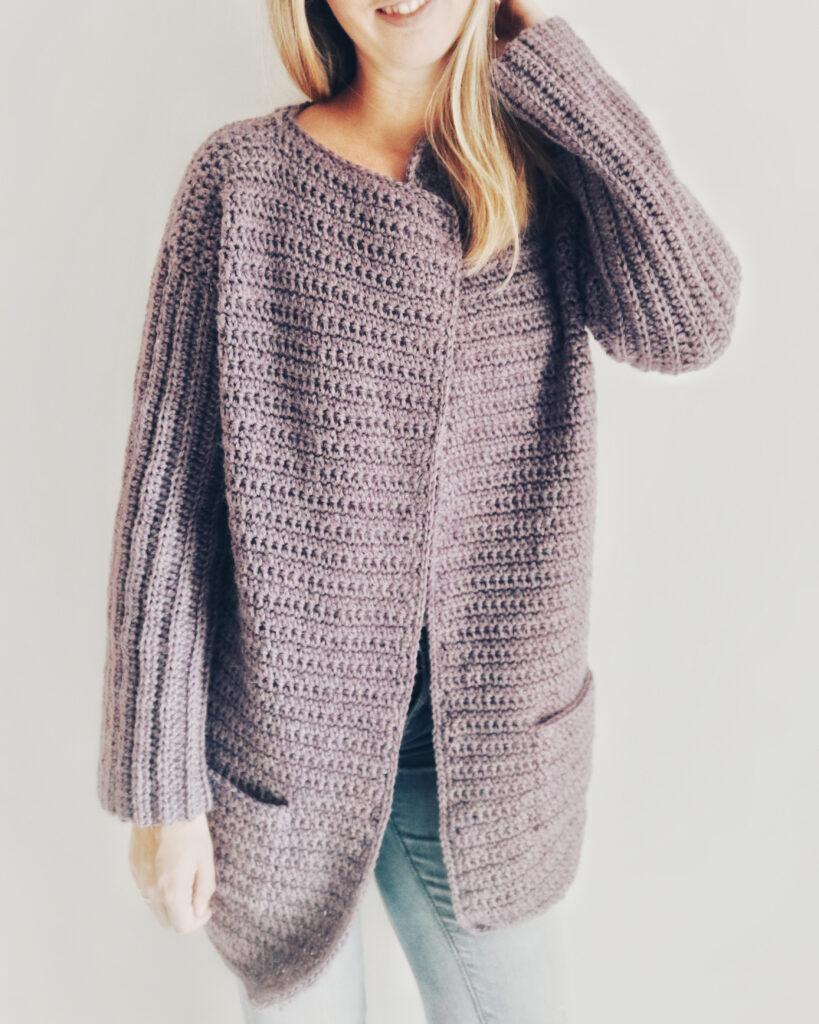 A woman wears a deep purple crochet cardigan and grey jeans. She is standing against a white background.