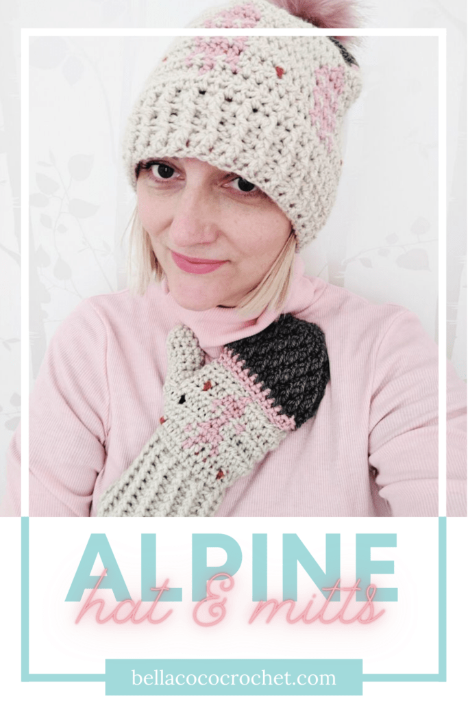 A woman wears a pink sweater and a crochet hat and mitten set. They are alpine-themed and in shades of tan, pink and grey.