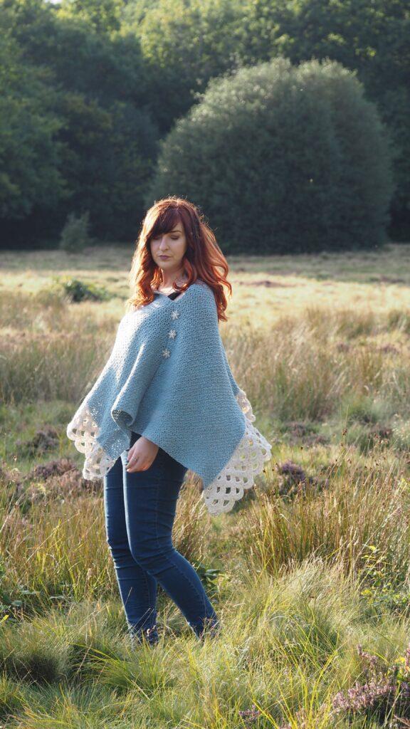 A woman with red hair is standing in moorland. She is wearing a crochet poncho in shades of blue and cream with a lace border.