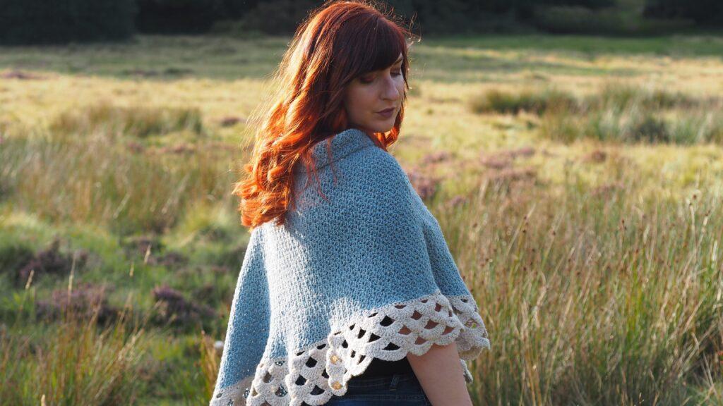 A woman with red hair is standing in moorland. She is wearing a crochet poncho in shades of blue and cream with a lace border.