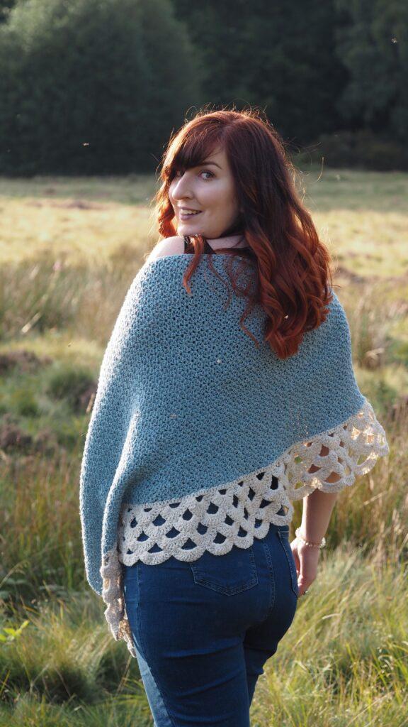 A woman with red hair is standing in moorland. She is wearing a crochet poncho in shades of blue and cream with a lace border.