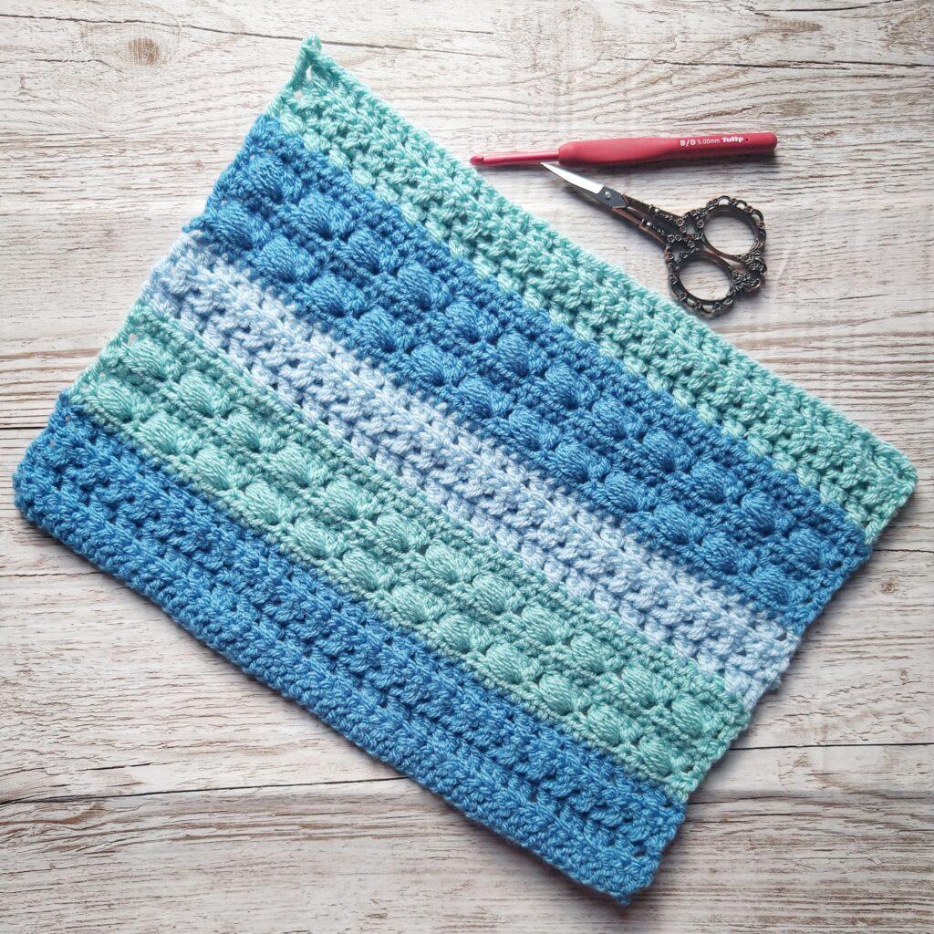 A blue crochet cushion in progress sits on a wooden background. The cushion has different colour and texture stripes in varying shades of blue. 