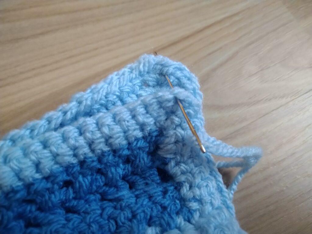 A blue crochet cushion in progress sits on a wooden background. The cushion has different colour and texture stripes in varying shades of blue.