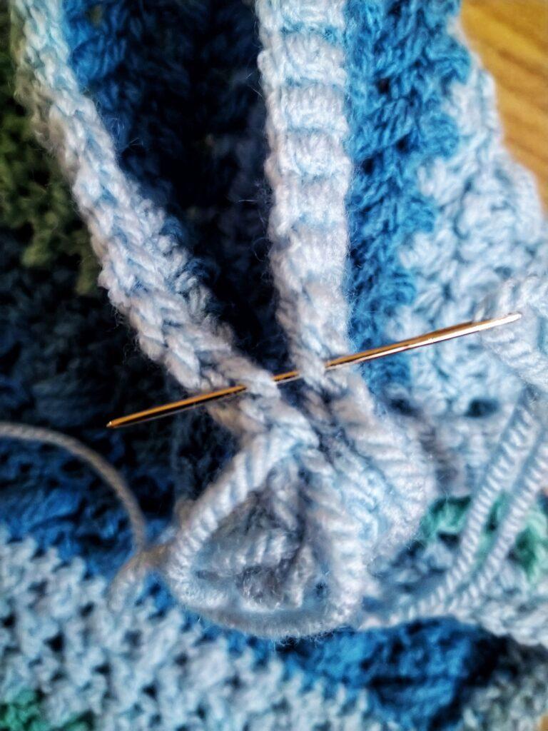 A blue crochet cushion in progress sits on a wooden background. The cushion has different colour and texture stripes in varying shades of blue.