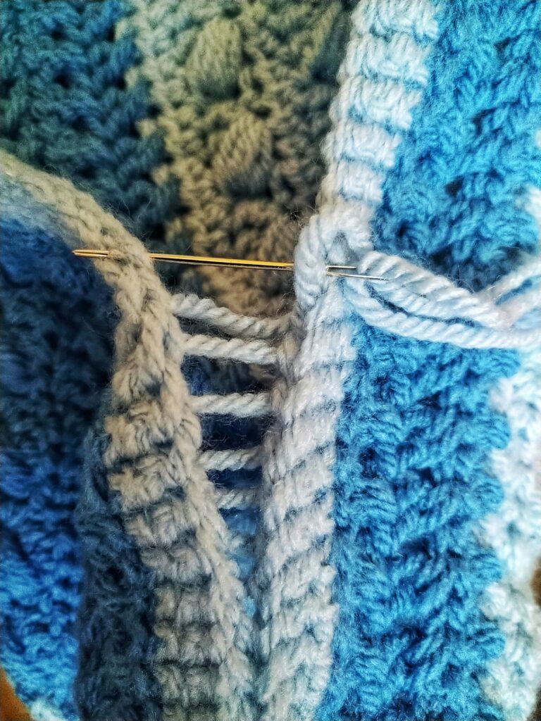 A blue crochet cushion in progress sits on a wooden background. The cushion has different colour and texture stripes in varying shades of blue.