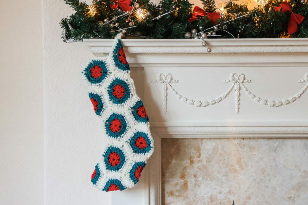 a crochet hexagon stocking made in red, green and cream yarn hanging from a cream mantle with a festive garland draped over the mantle piece.