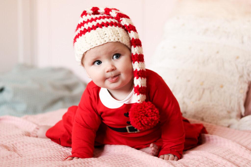 baby sat on a bed with pale pink bedding wearing a red Christmas dress and a crochet baby elf hat.
