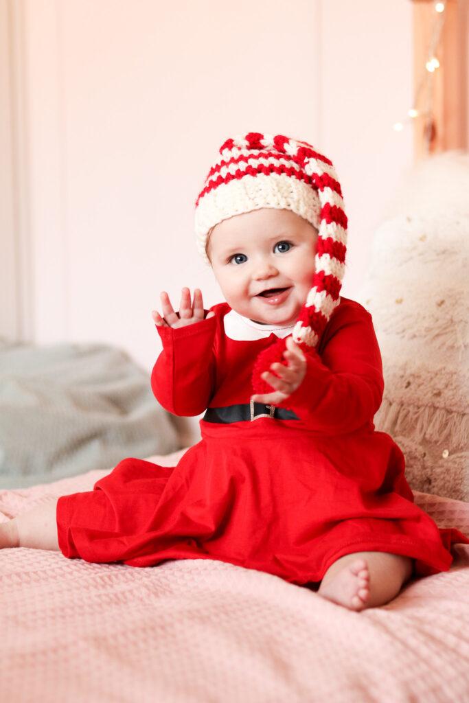 baby sat on a bed with pale pink bedding wearing a red Christmas dress and a crochet baby elf hat.