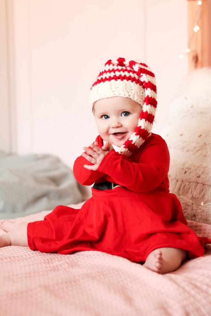 baby sat on a bed with pale pink bedding wearing a red Christmas dress and a crochet baby elf hat.