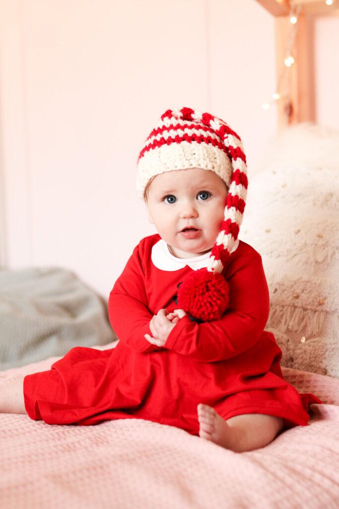 baby sat on a bed with pale pink bedding wearing a red Christmas dress and a crochet baby elf hat.