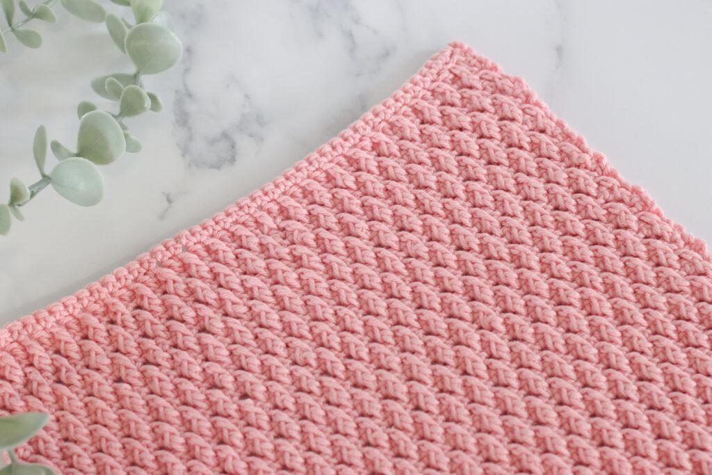 a pink crochet swatch of the alpine stitch on a marbled backdrop and foliage to the left of the image.