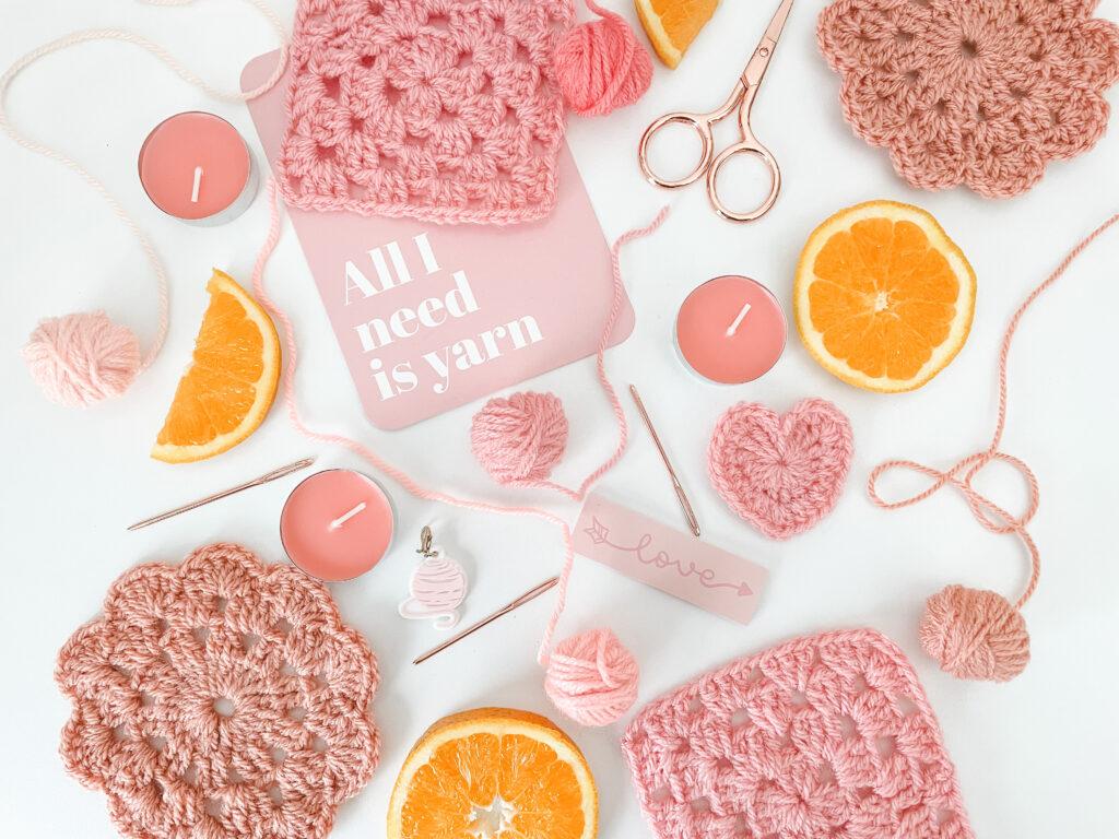 a flay lay of pink objects including, a crochet coaster and granny square, yarn, candles, rose gold scissors, darning needle and pink tea light candles.