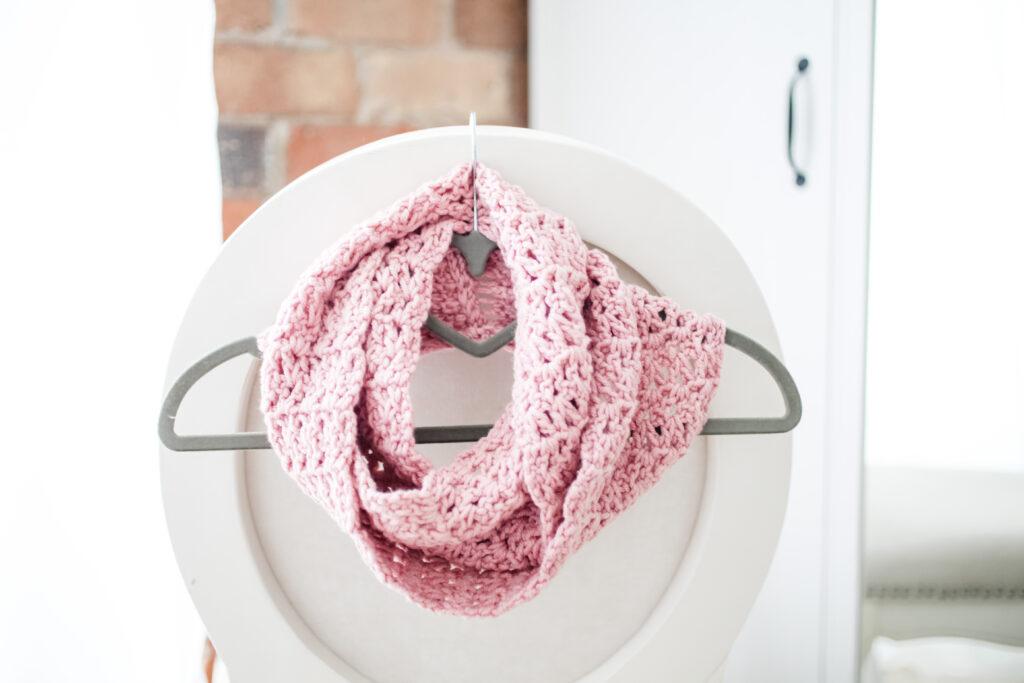 a pink infinity scarf wrapped around a grey clothes hanger thats hooked on the back of a white chair.