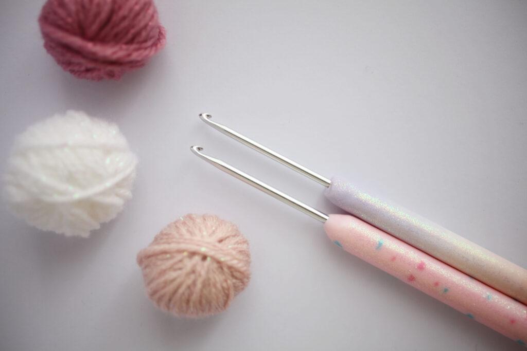 A pair of crochet hooks with sparkly handles sit on a white background alongside three small balls of yarn in shades of pink.