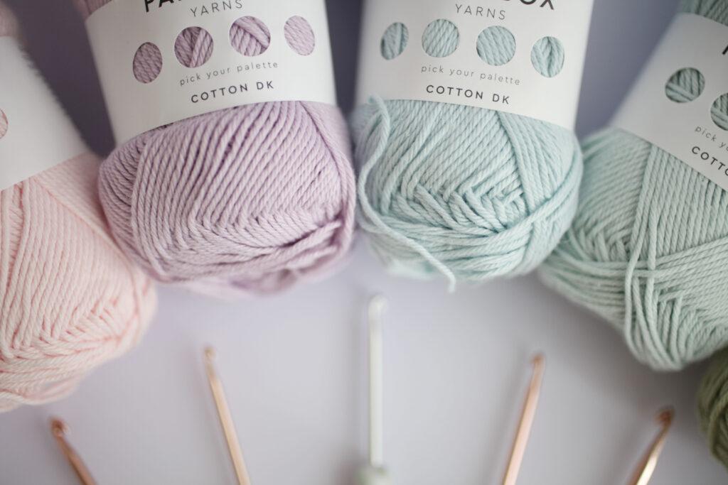 Four balls of yarn are sat on a white backdrop with hooks faintly visible in the background.